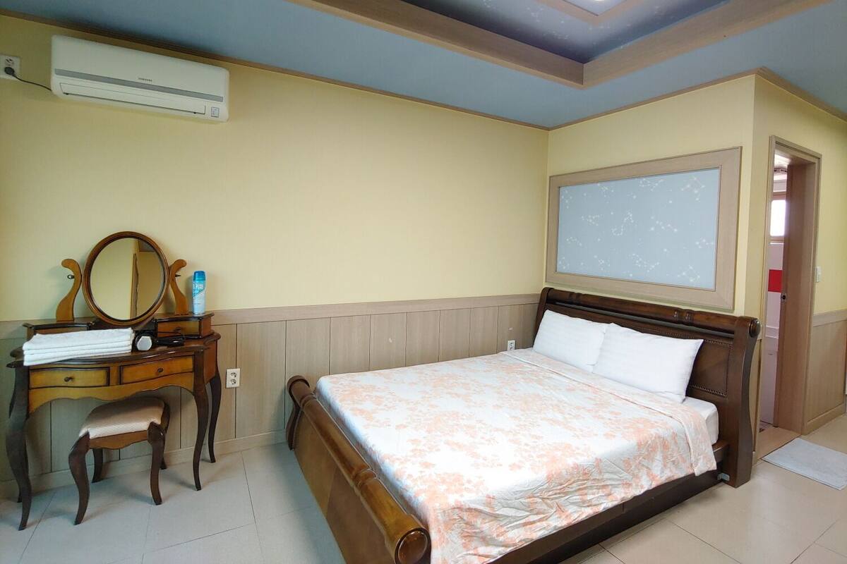 Un dormitorio con cama, un escritorio de madera con espejo, aire acondicionado de pared y ventana con vista al cielo.