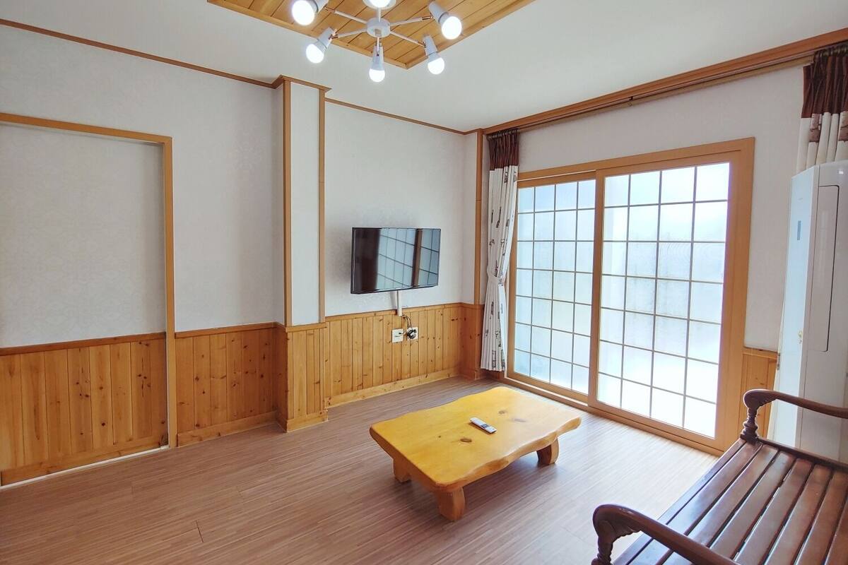 Una habitación con paneles de madera, un ventilador de techo, un televisor montado en la pared, una ventana con cortinas y un banco de madera.