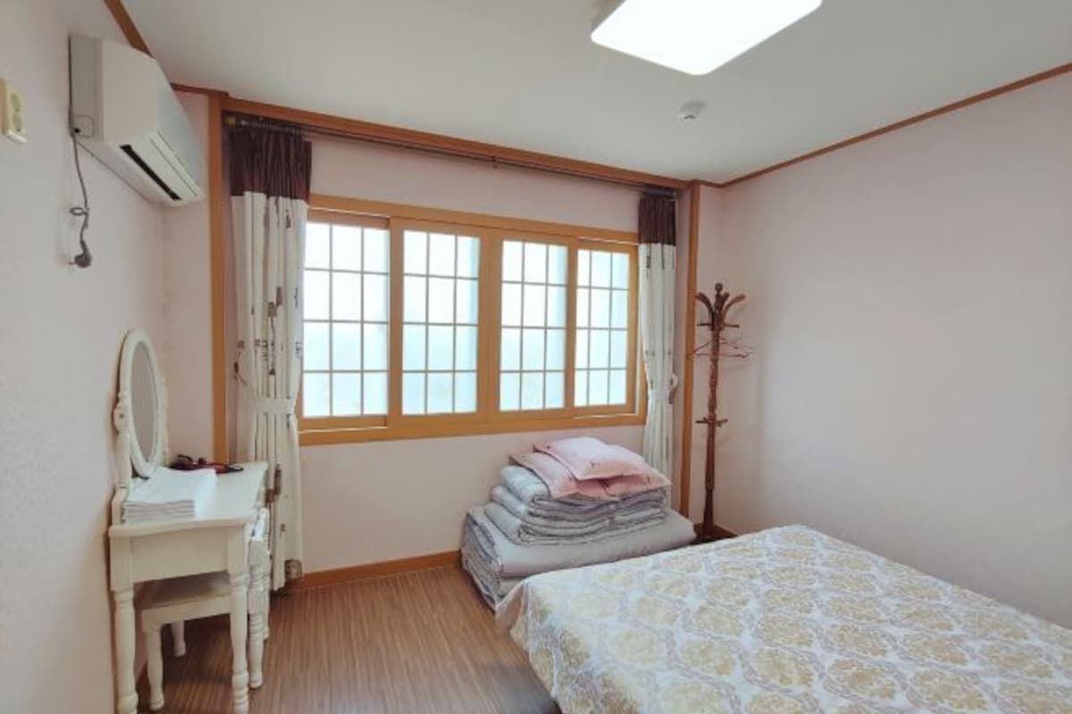 A bedroom with a bed, a desk, and a chair. There is a window with curtains and a coat rack.