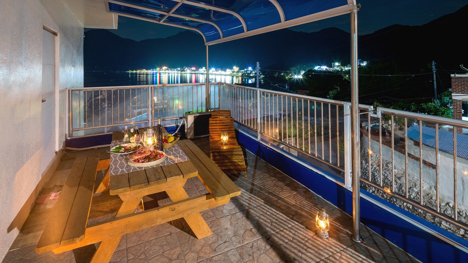 A balcony with a wooden table set for dining, a railing, and a view of a cityscape at night.