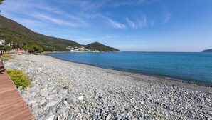 A pebble beach with a wooden walkway, calm sea, and hills in the background. - Melodia Pension (Geoje)