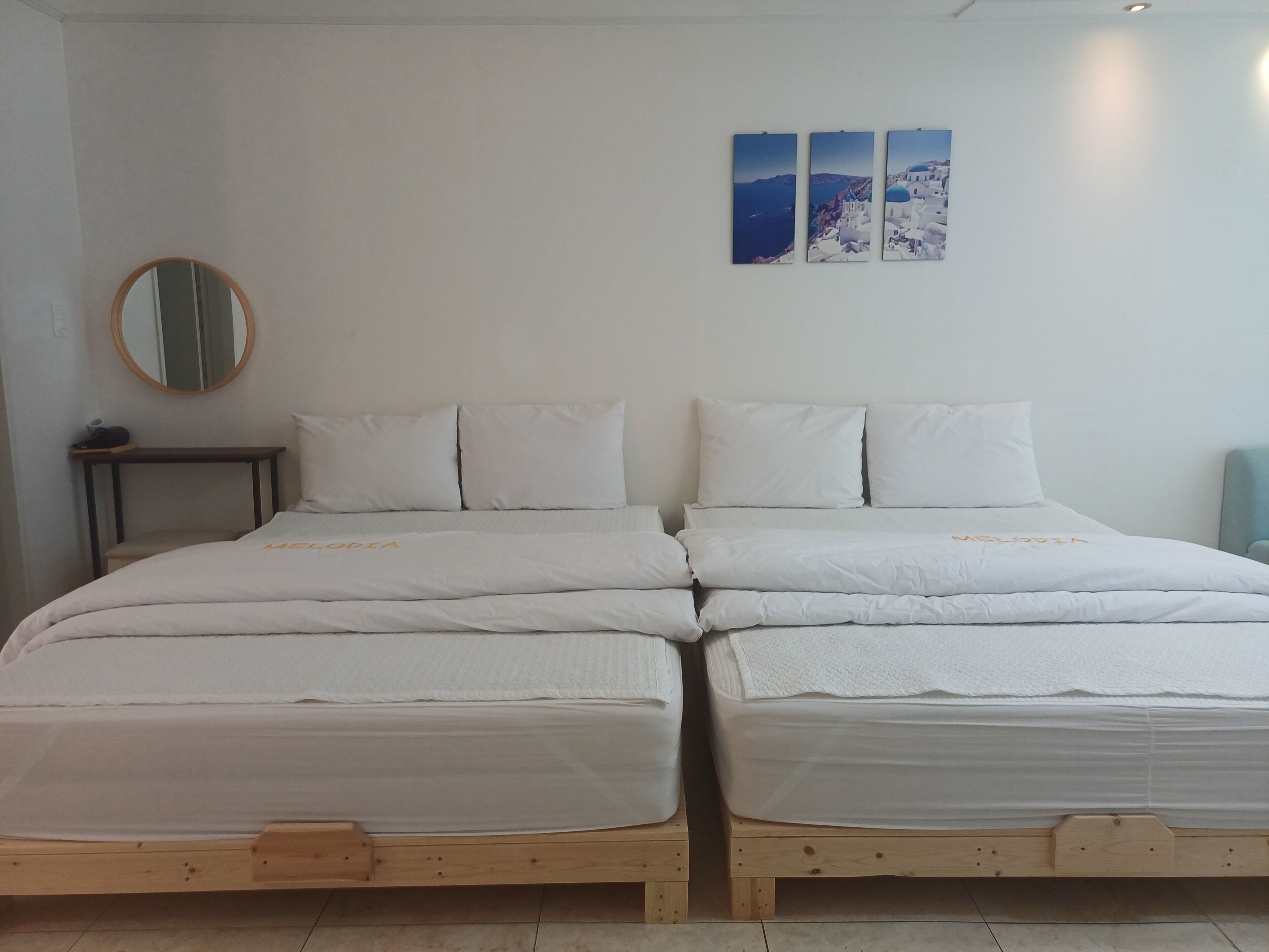 Cama de casal com estrutura de madeira e roupa de cama branca, mesa de cabeceira com abajur e espelho fixado na parede.
