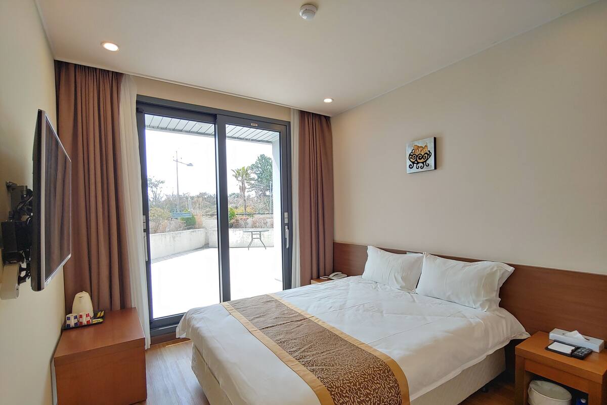 Quarto de hotel com uma cama grande, mesa de cabeceira com telefone e lenços, uma lixeira pequena e uma televisão na parede.