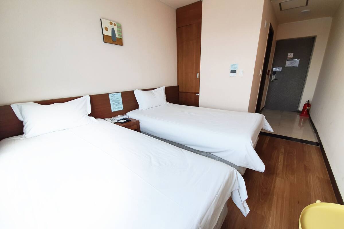 Quarto de hotel com duas camas, cabeceira de madeira, mesa de cabeceira pequena com telefone e uma cadeira amarela.