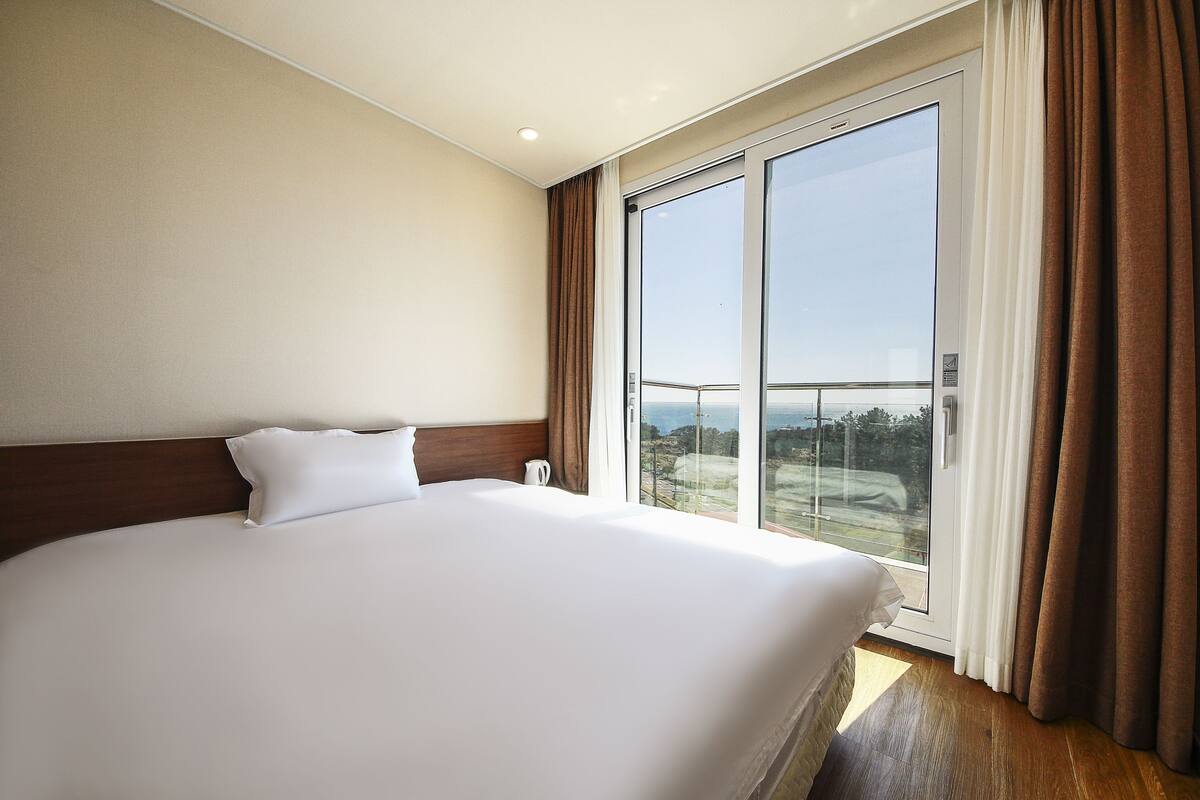 Quarto de hotel com uma cama grande, vista através de uma porta de correr de vidro e piso de madeira.
