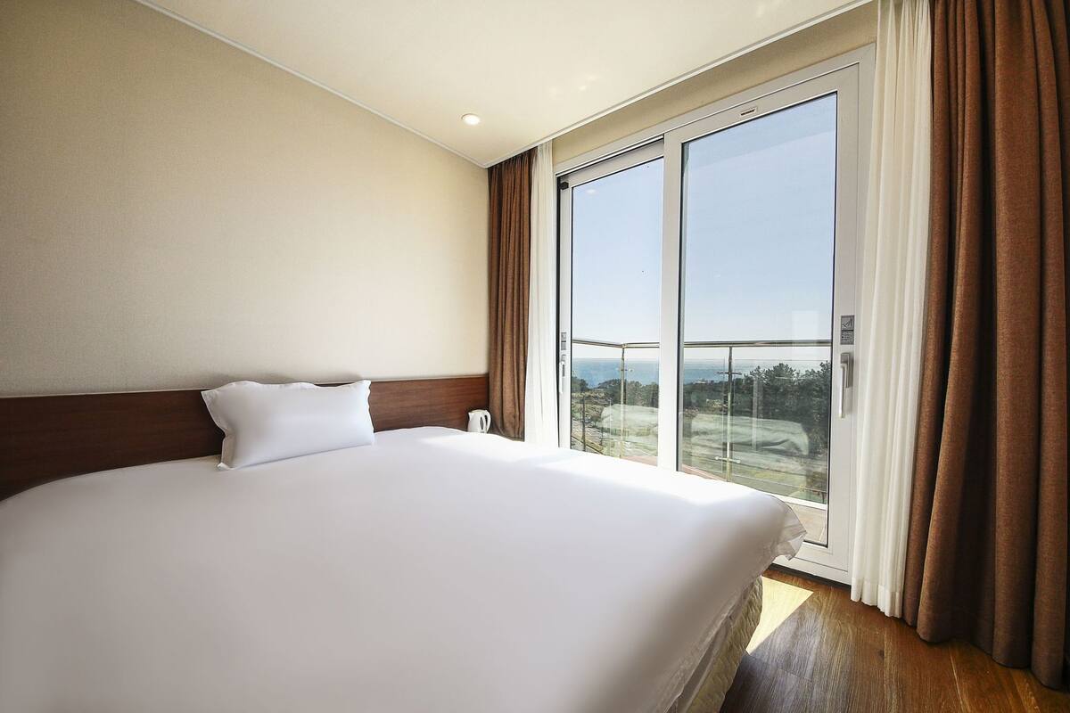 Quarto de hotel com uma cama grande, vista através de uma porta de correr de vidro e piso de madeira.