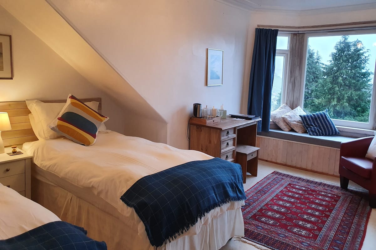 Un dormitorio con cama, escritorio, silla, ventana con cortinas y vistas a árboles.