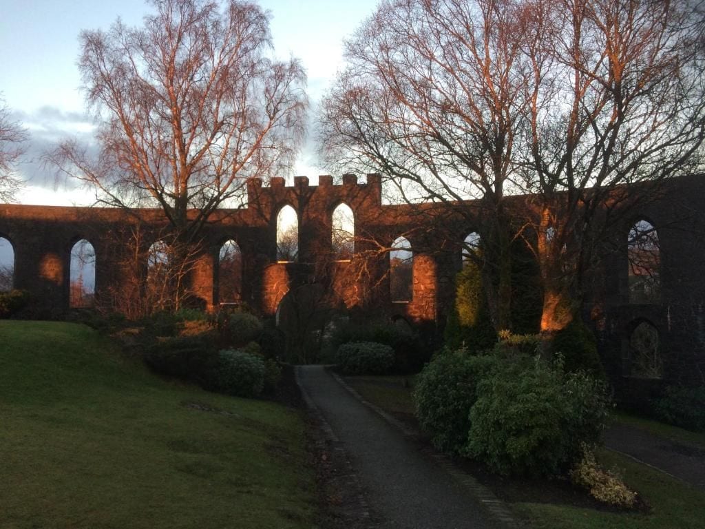 Un puente de piedra histórico con pasarelas en arco, rodeado de árboles y un césped bien cuidado.