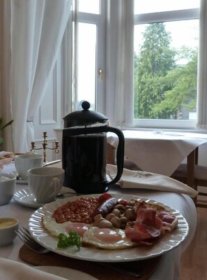 Bandeja de desayuno con huevos, frijoles, salchichas y tocino en un plato, una cafetera y una taza sobre una mesa junto a una ventana.
