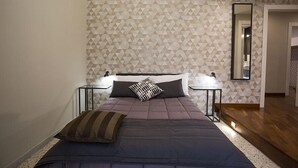 A bedroom with a geometric patterned wall, a bed with a dark blue bedspread and two pillows, a nightstand with a lamp, and a mirror on the wall. - B&B Rettifilo (Naples)