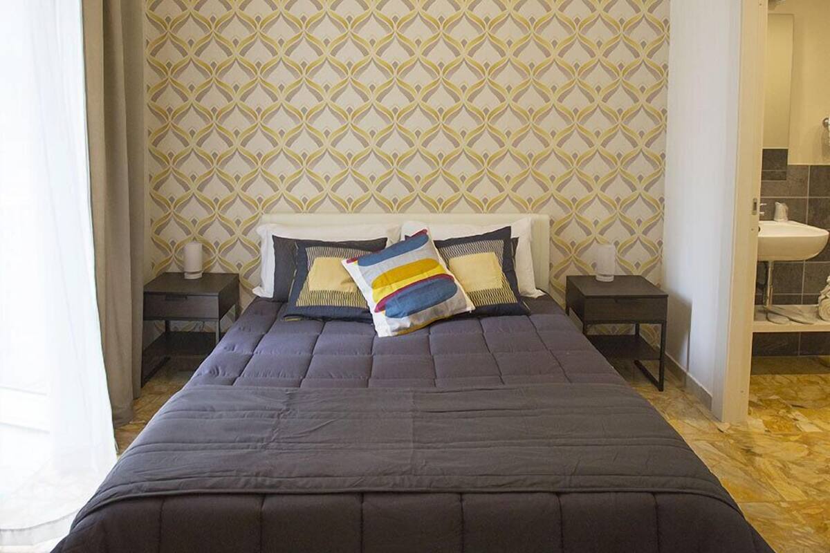 Cama bem arrumada com colcha escura, duas mesas de cabeceira com luminárias e papel de parede estampado.