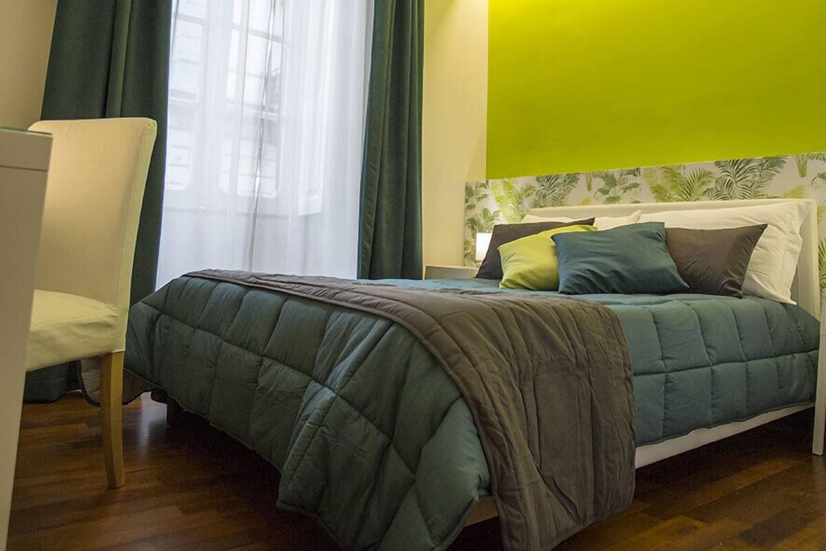 Quarto com cama, cadeira, janela com cortinas e uma parede verde de destaque.