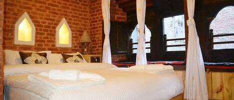 A bed with white bedding, a wooden headboard, and a patterned rug. There are two windows with white curtains and a brick wall.