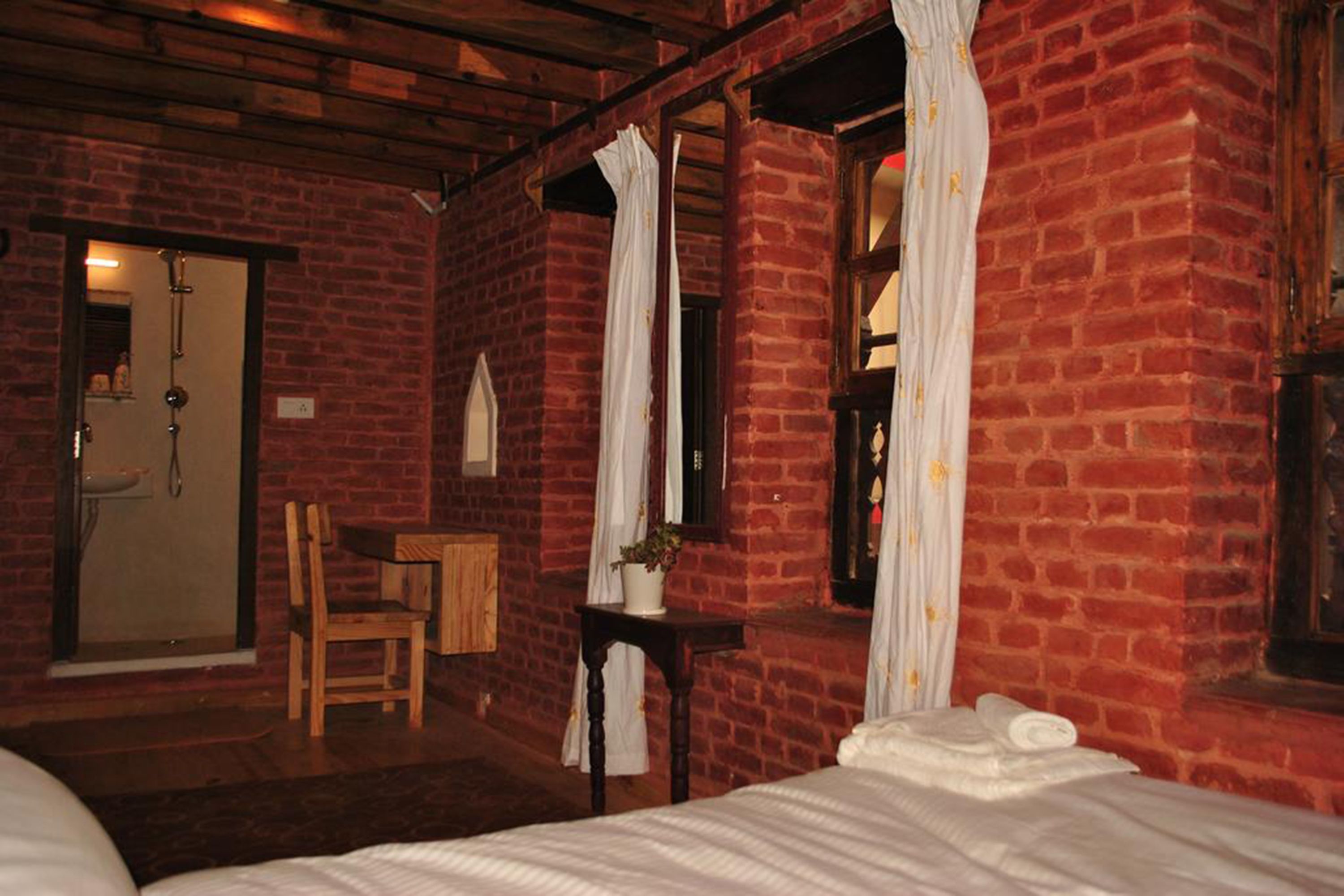 Ein Zimmer mit Backsteinwand, einem Bett, einem Sessel, einem kleinen Tisch und einem Fenster mit Vorhängen.