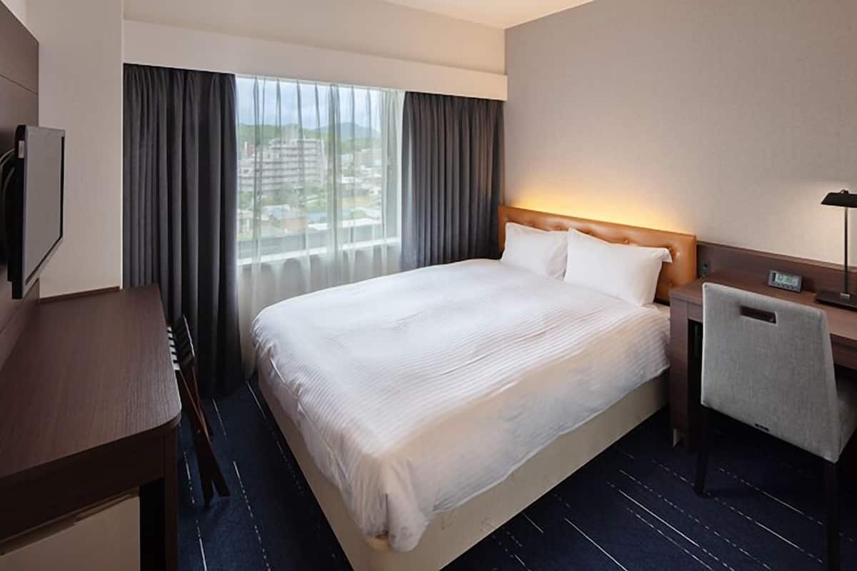 A hotel room with a large bed, a desk, a chair, a nightstand, and a view of the city through the window.