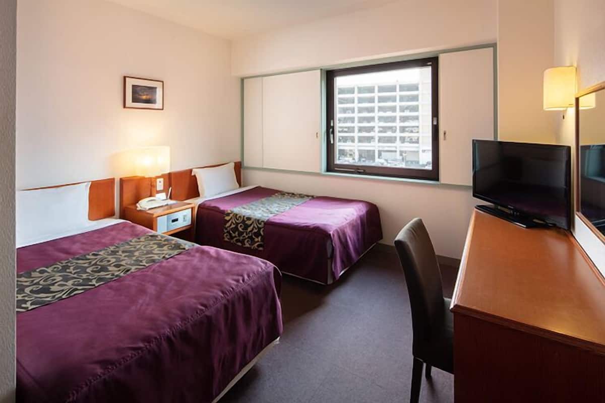 A hotel room with two beds, a desk, a television, and a window with a view of buildings.