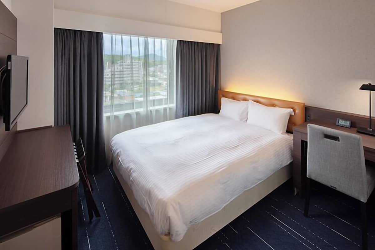 A hotel room with a large bed, a desk, a chair, a nightstand, and a view of the city through the window.