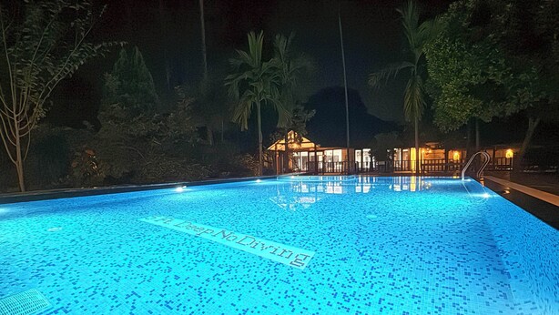 Une piscine éclairée la nuit, avec un sol carrelé et un bâtiment visible à l’arrière-plan.