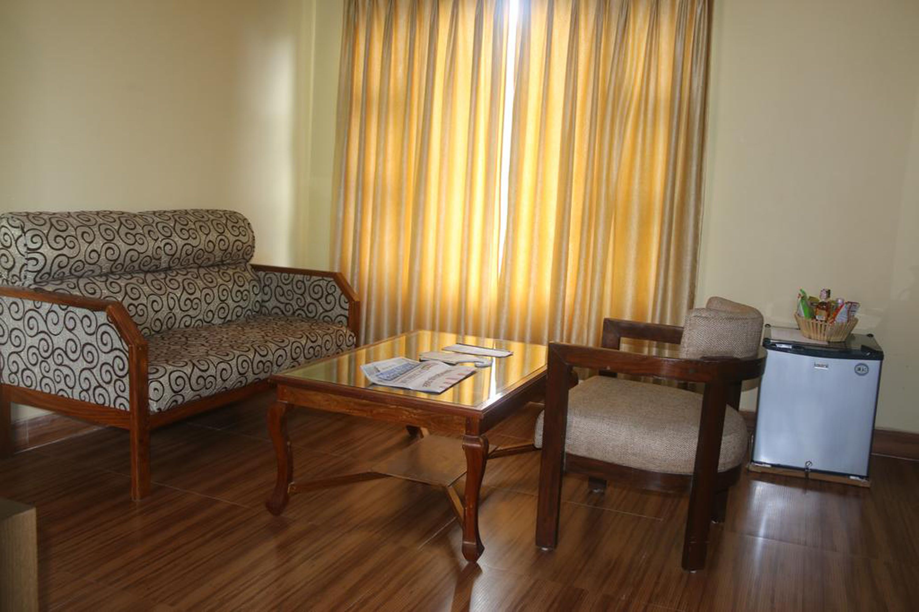 A living room with a patterned sofa, a wooden coffee table, a small table with a basket, and a compact refrigerator.