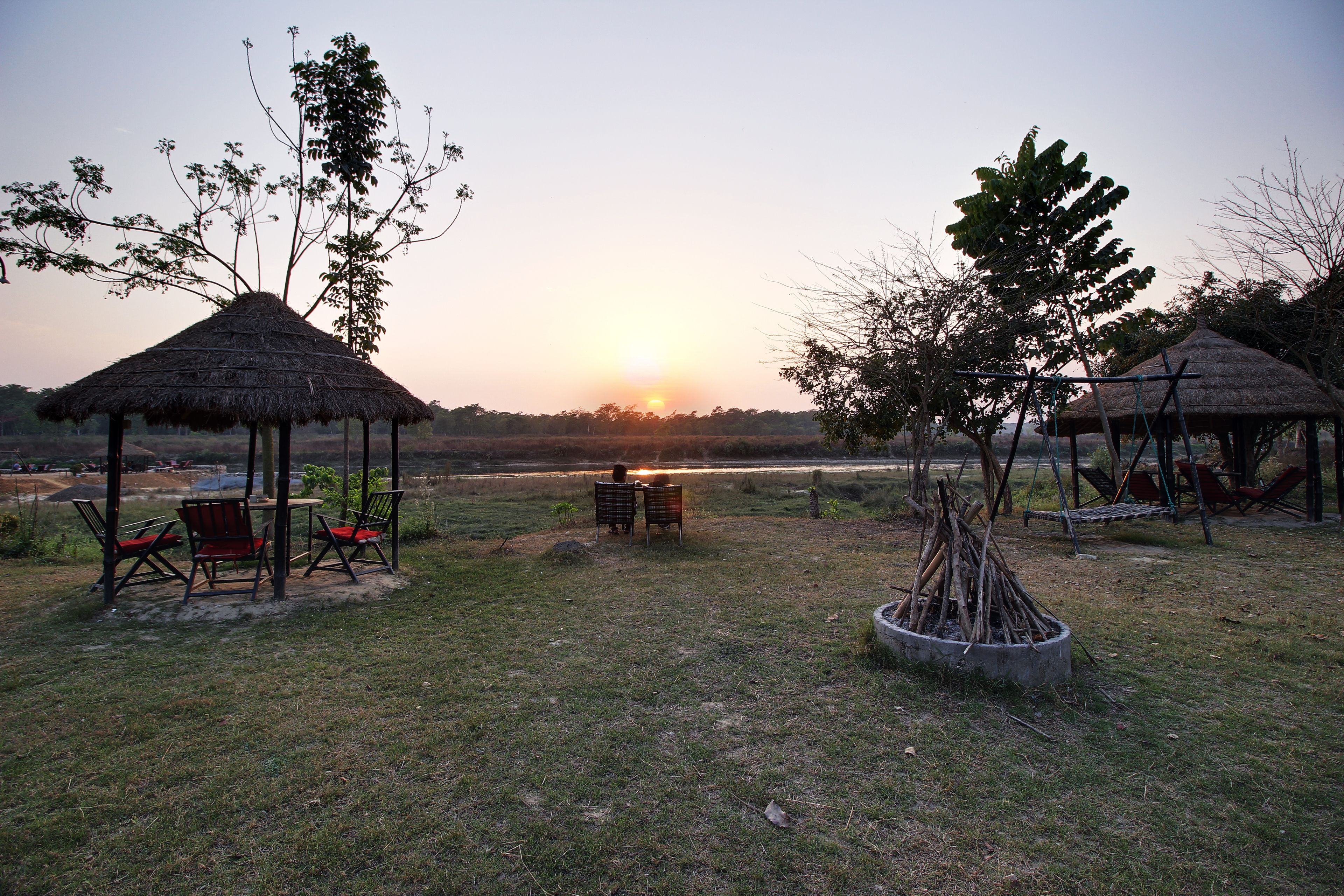 A thatched-roof pavilion with outdoor seating, surrounded by trees and a grassy area.