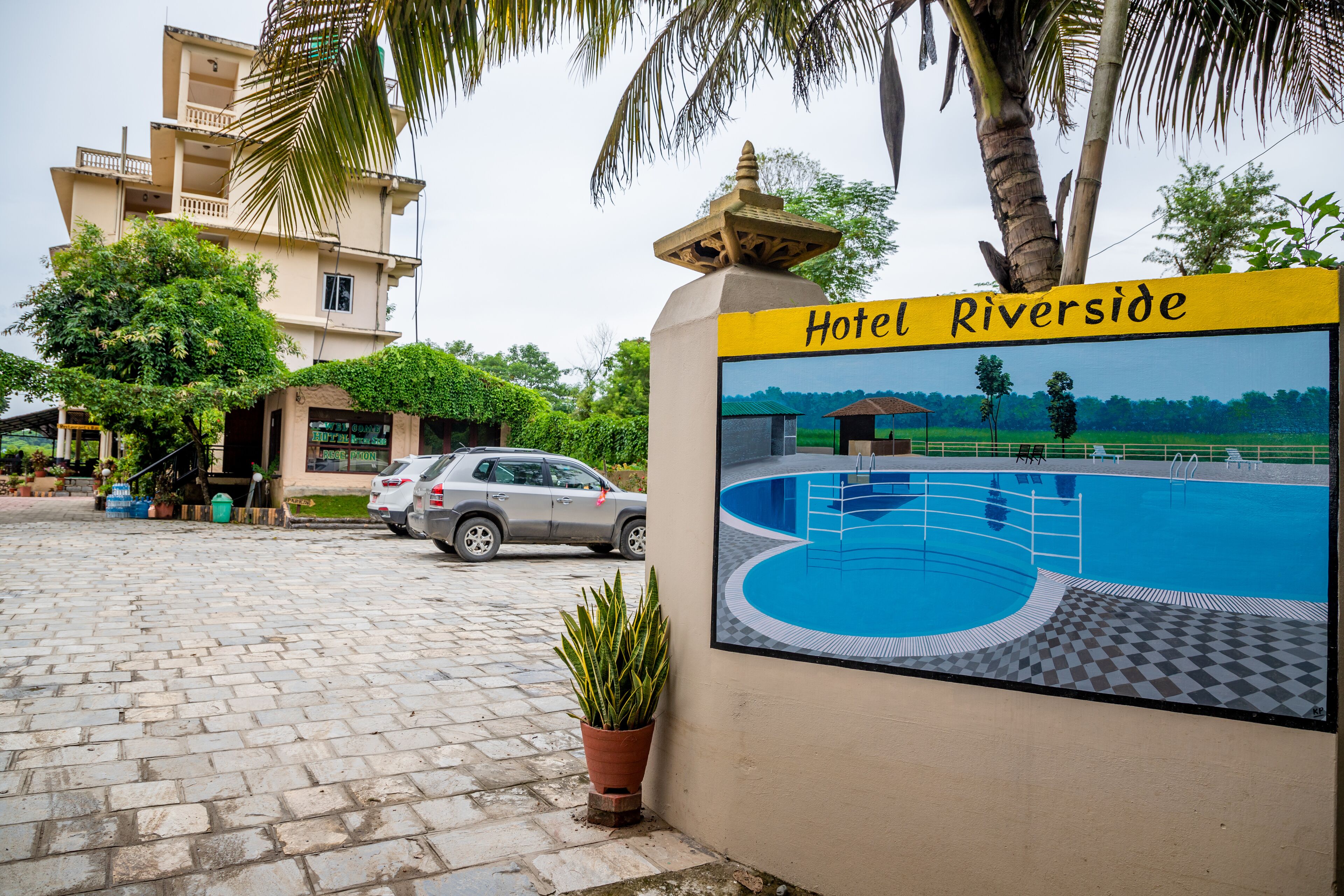 A hotel entrance with a sign reading "Hotel Riverside," a paved driveway, a parked car, and a pool area depicted in the background.