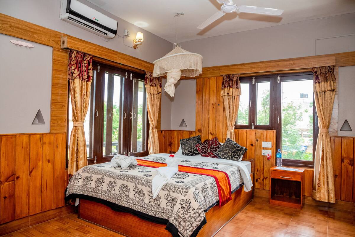 A bedroom with a wooden floor, a bed with patterned bedding, a bedside table, a nightstand, a hanging mosquito net, and a window with curtains.