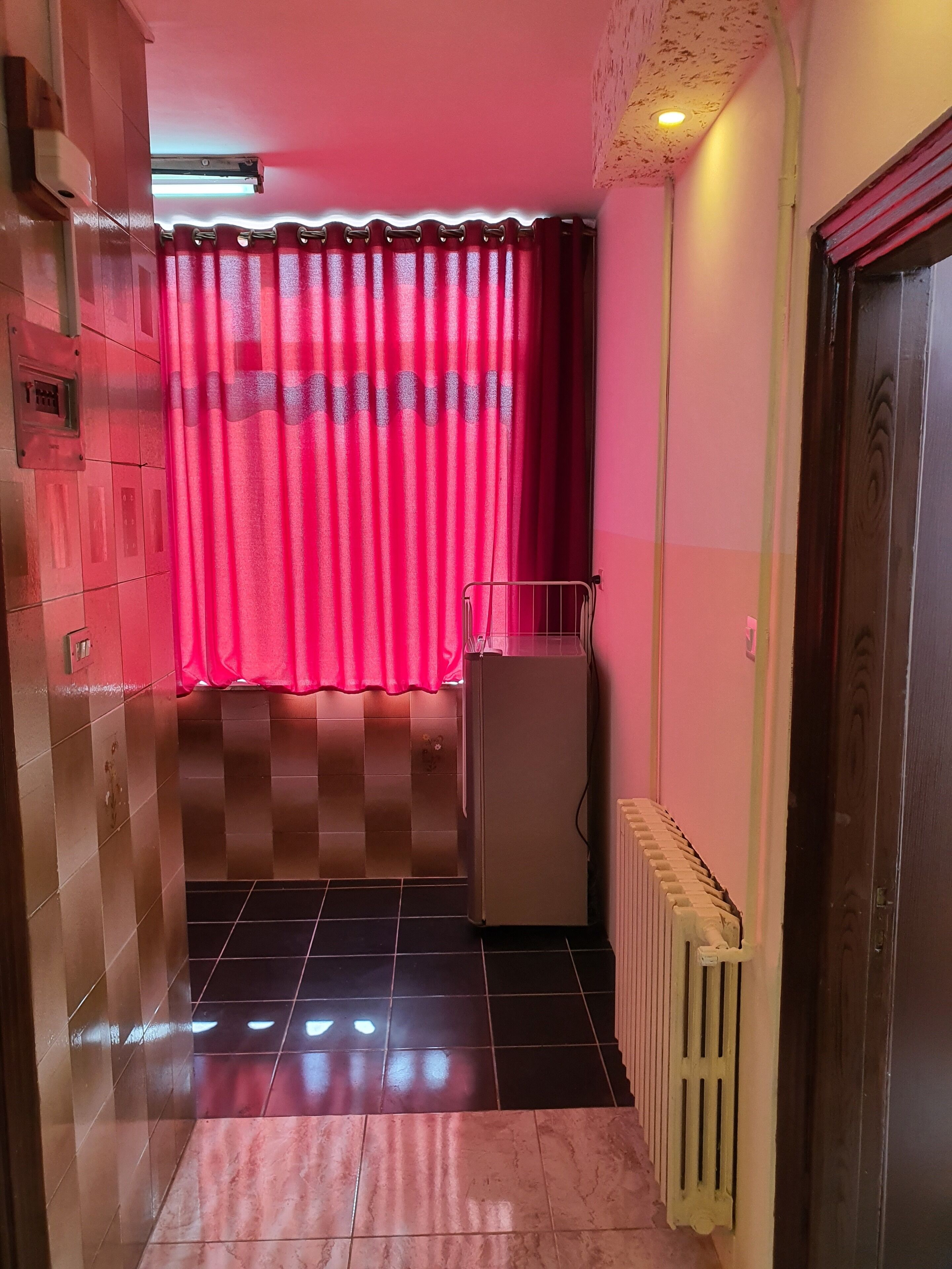 Een smalle gang met een betegelde vloer, een radiator, een koelkast en een deur aan de rechterkant.
