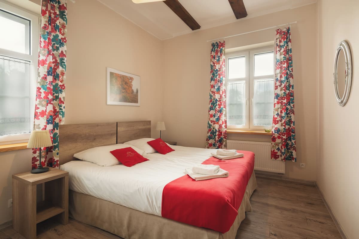 A neatly made bed with red bedding, a bedside table with a lamp, and floral curtains on the windows.