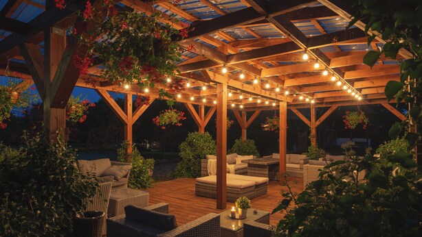 Un espacio de patio al aire libre con pérgola de madera, mobiliario de mimbre e iluminación con luces colgantes.