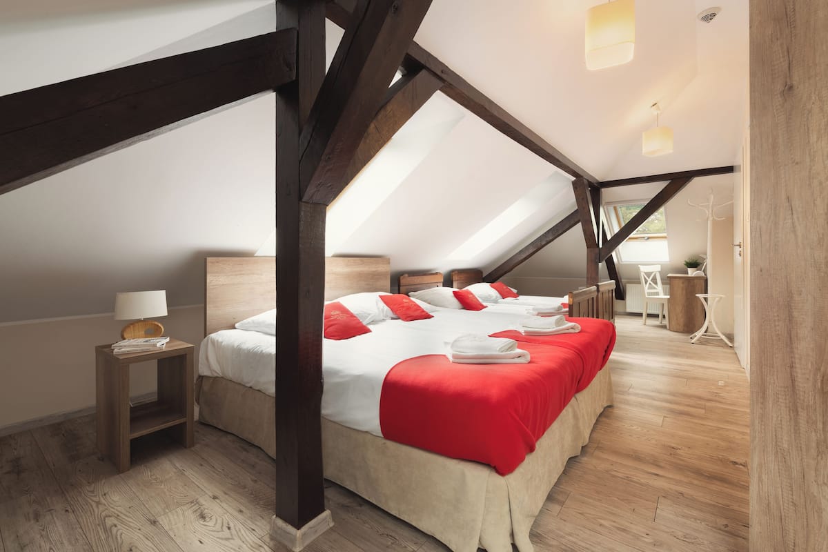 A bedroom with a sloped ceiling, wooden beams, a large bed with red bedding, a bedside table with a lamp, and a view of the room.