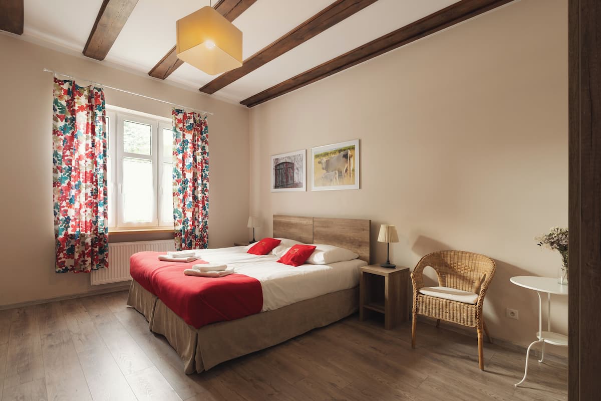 A bedroom with a wooden floor, a bed with red bedding, a wicker chair, and a small table with a lamp.