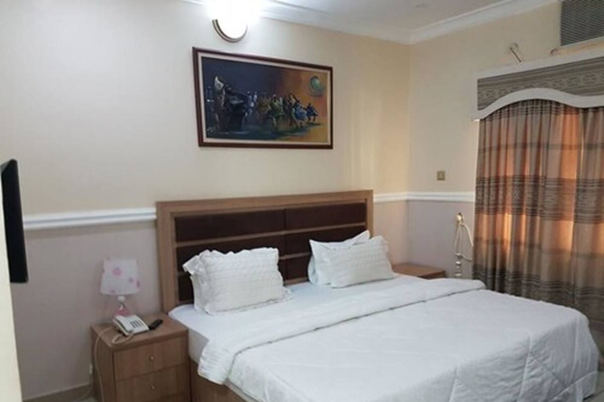 A hotel room with a large bed, a nightstand with a lamp, a painting on the wall, and a window with curtains.