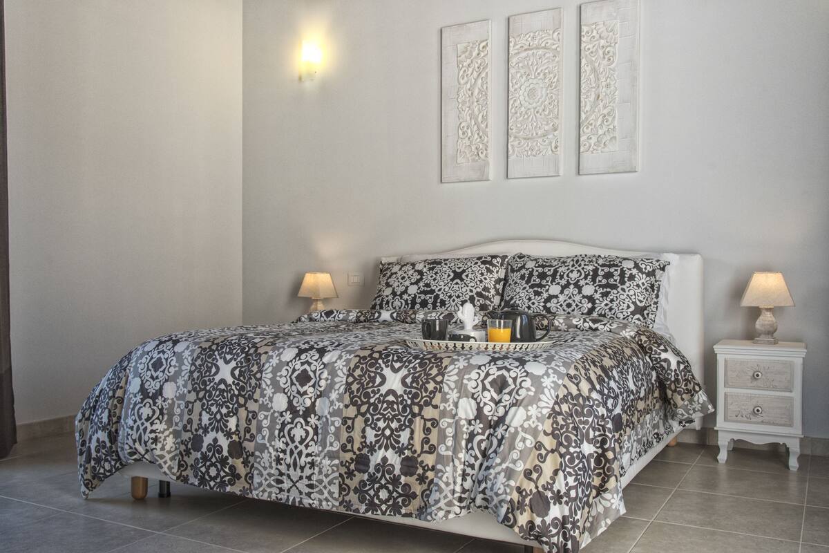 A neatly made bed with a patterned bedspread, two bedside lamps, and a tray with a cup and a book on the nightstand.