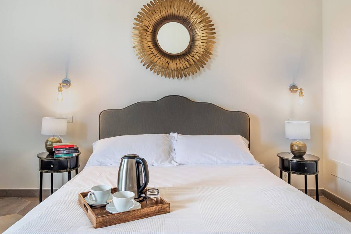 A neatly made bed with white linens, a wooden tray with cups and a kettle, and a decorative mirror on the wall.