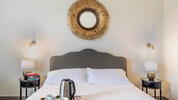 Un lit bien fait, avec du linge de lit blanc, un plateau en bois avec des tasses et une bouilloire, et un miroir décoratif accroché au mur.