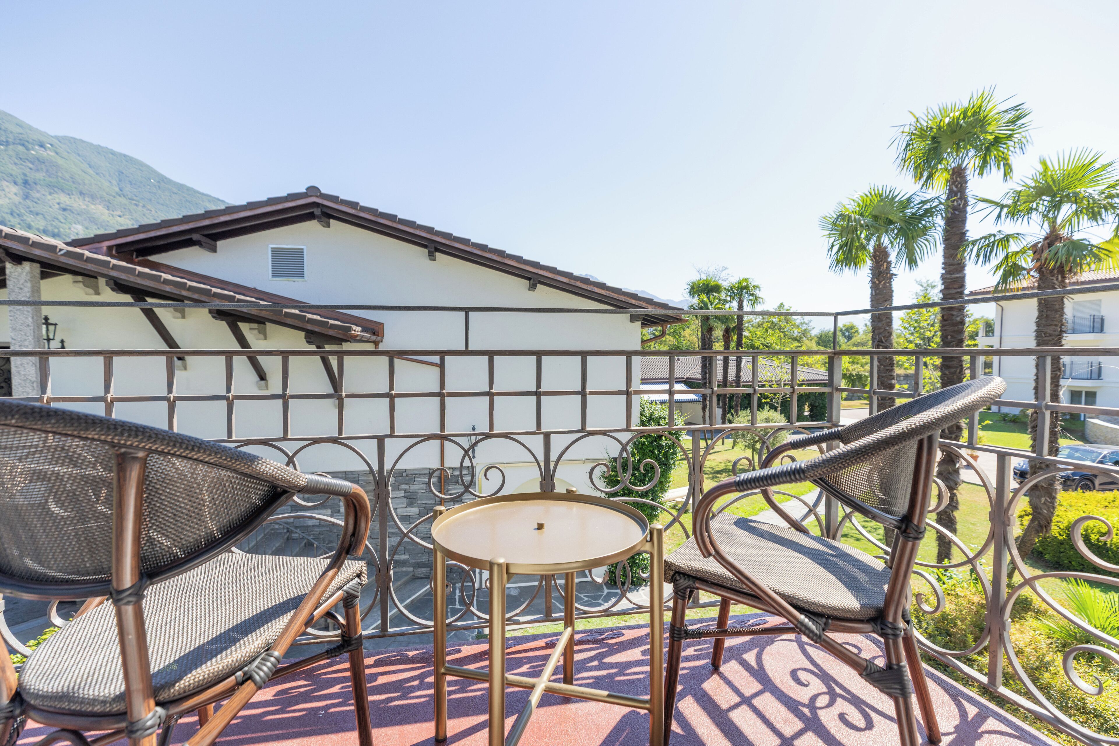 A balcony with wicker chairs and a small table, overlooking a garden with palm trees and a building in the background.