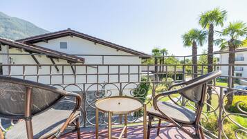 A balcony with wicker chairs and a small table, overlooking a garden with palm trees and a building in the background.