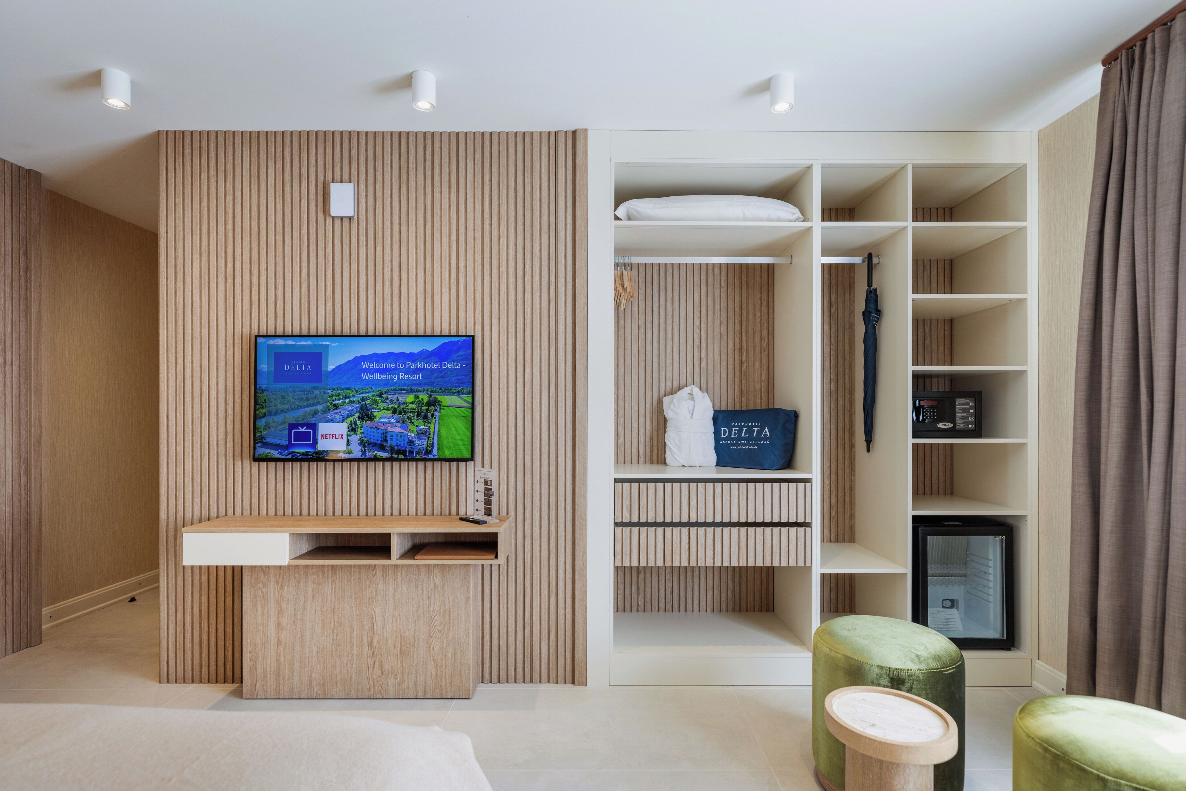 Una sala moderna con un televisor de pantalla plana montado en una pared de madera, un mueble empotrado para entretenimiento, una zona de estar con otomanas y un vestidor con estantes y espacio para colgar.