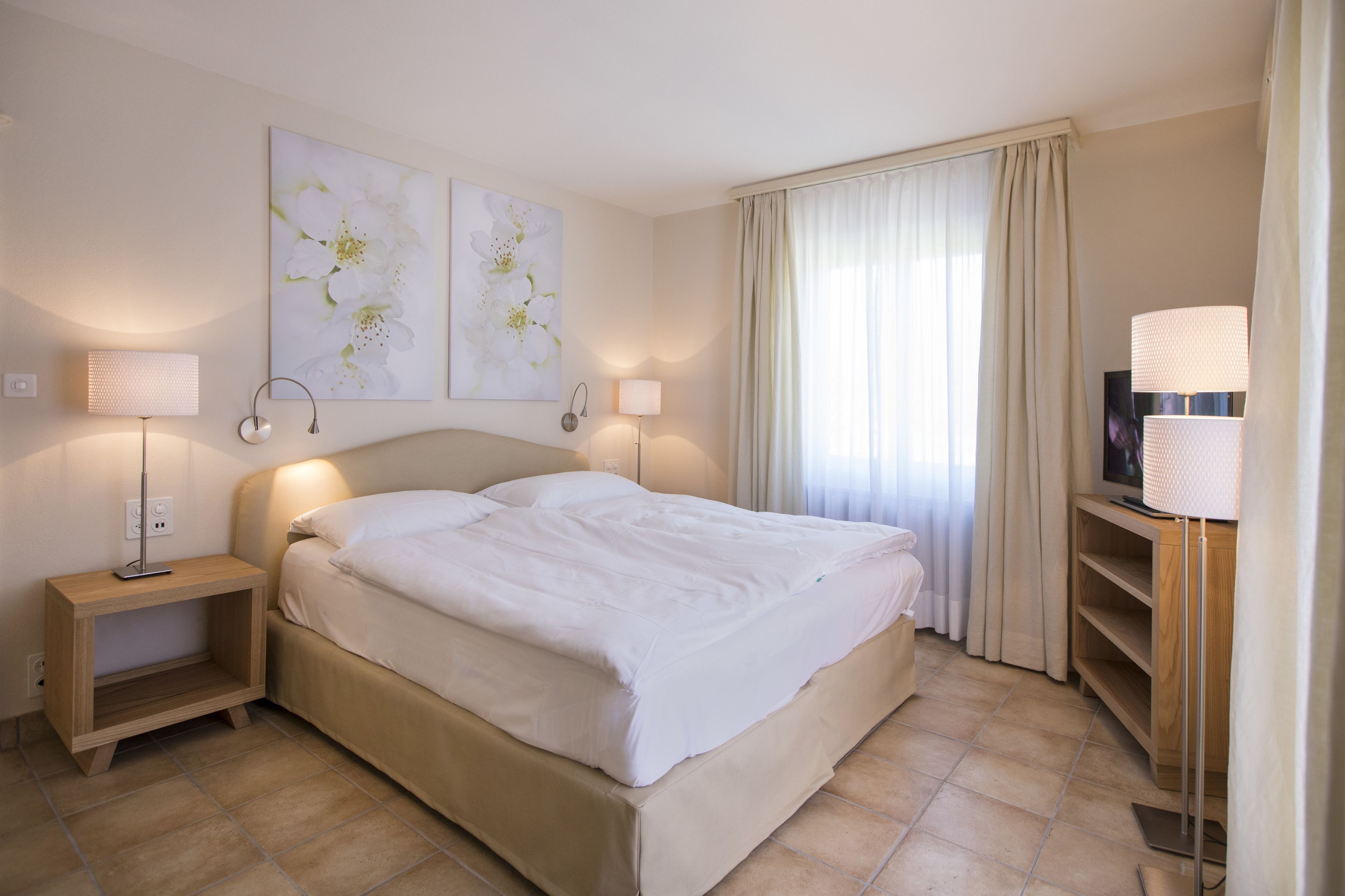 Habitación de hotel ordenada con una cama, dos lámparas de noche, un televisor sobre un mueble bajo y un ventanal con cortinas.