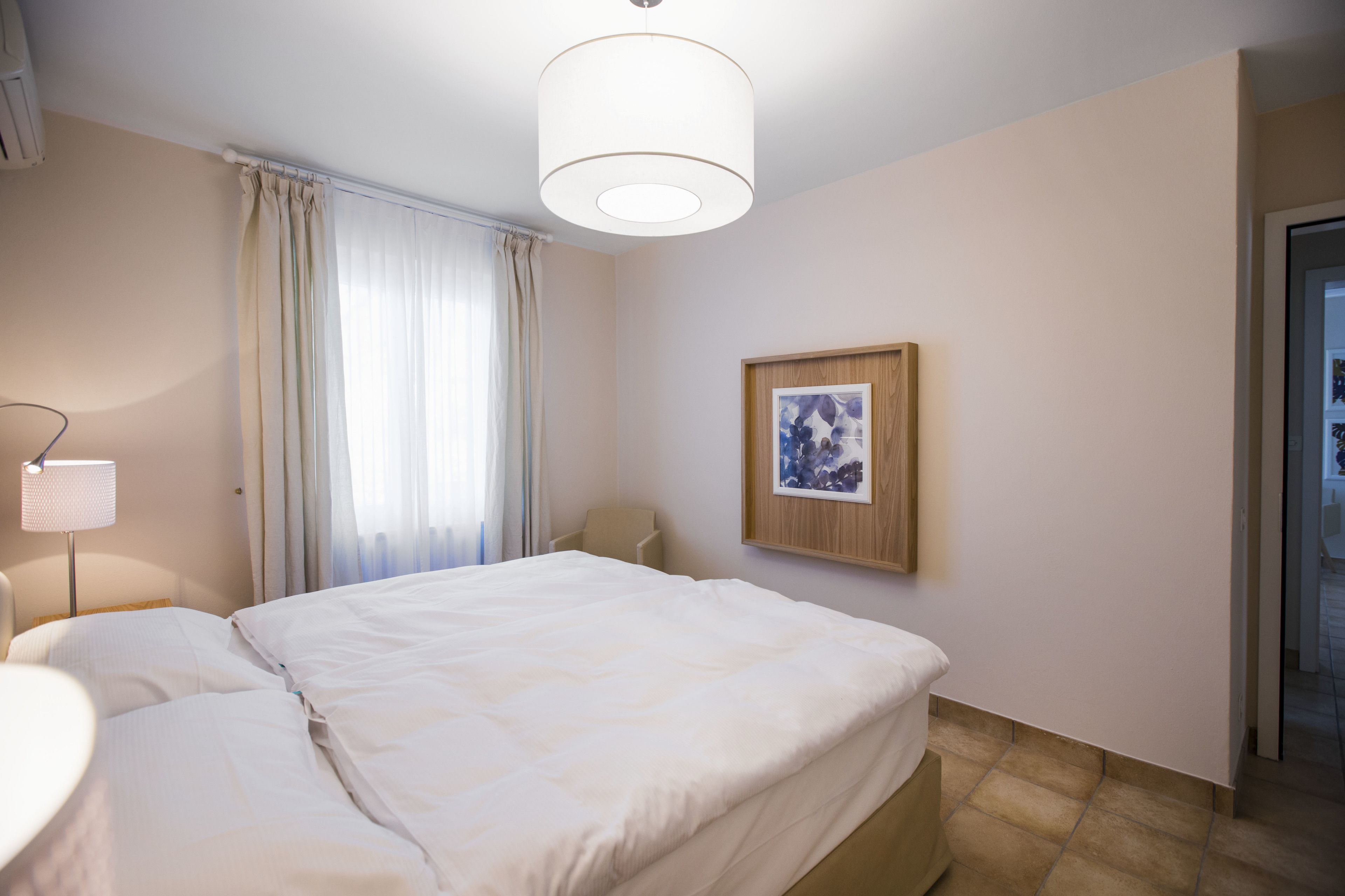 Una camera d'hotel con un letto, un comodino, una lampada, un quadro appeso al muro e una porta che dà su un'altra stanza.