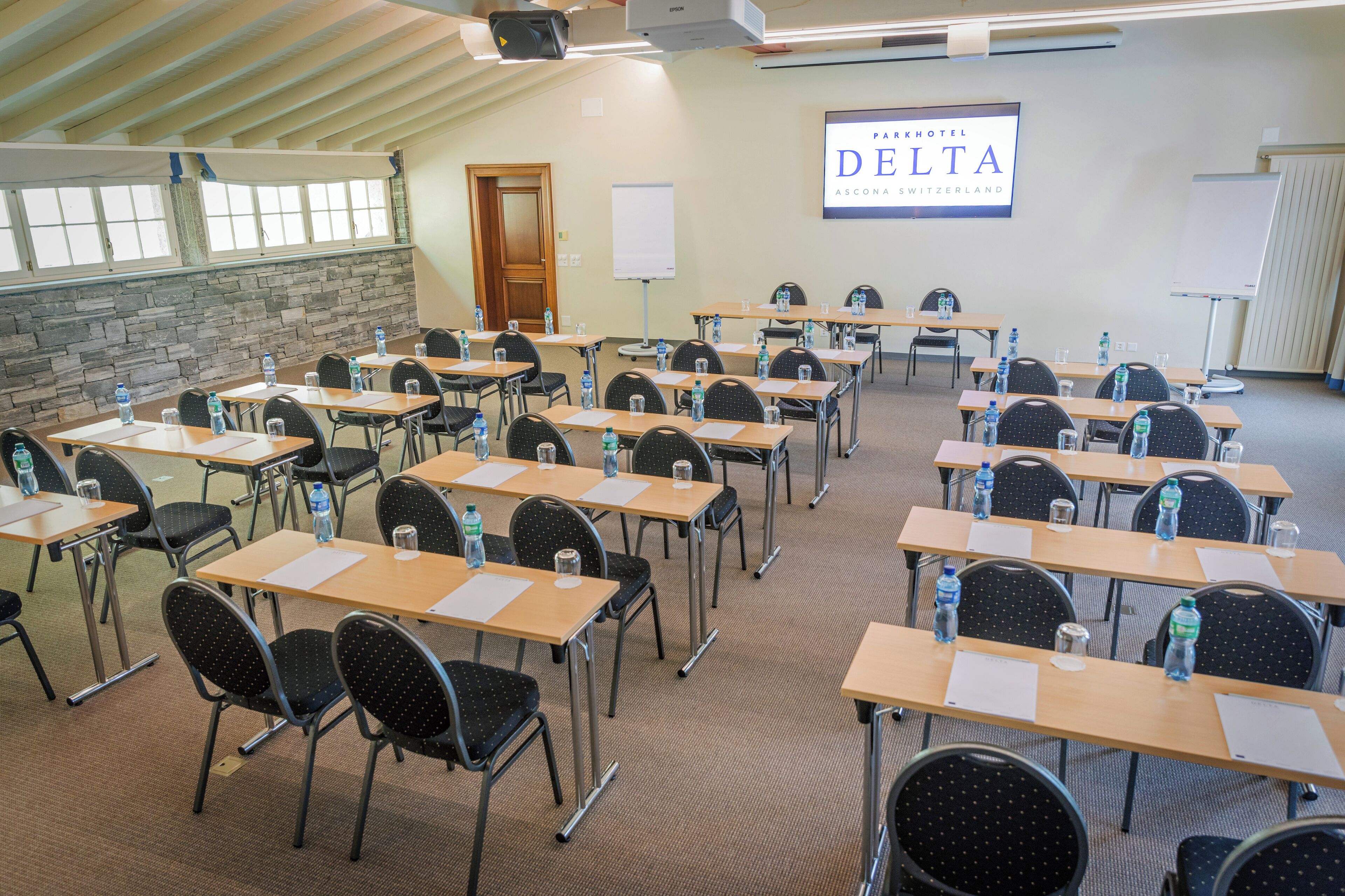 Un salón de conferencias con filas de mesas y sillas, una pantalla de proyector y un letrero que dice Parkhotel Delta.