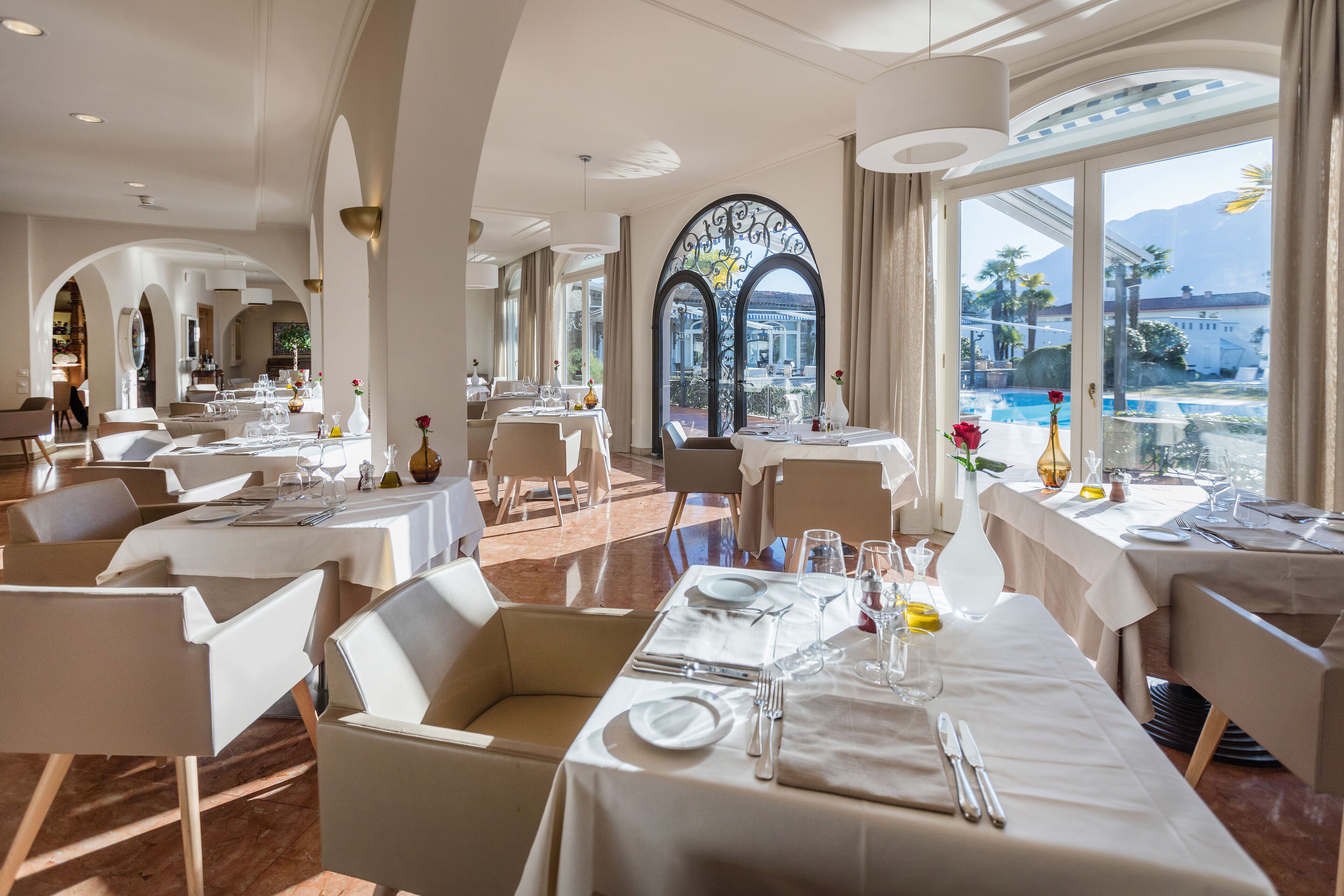 A spacious dining area with multiple tables set for dining, large windows offering a view of a pool and mountains, and a modern architectural design with arched ceilings and large windows.