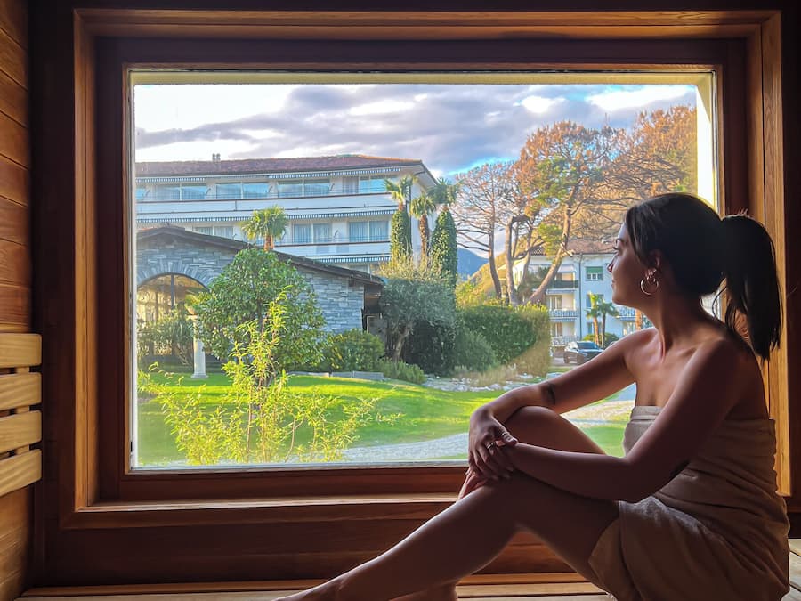 Una persona sentada junto a una ventana con vistas a un edificio y vegetación.