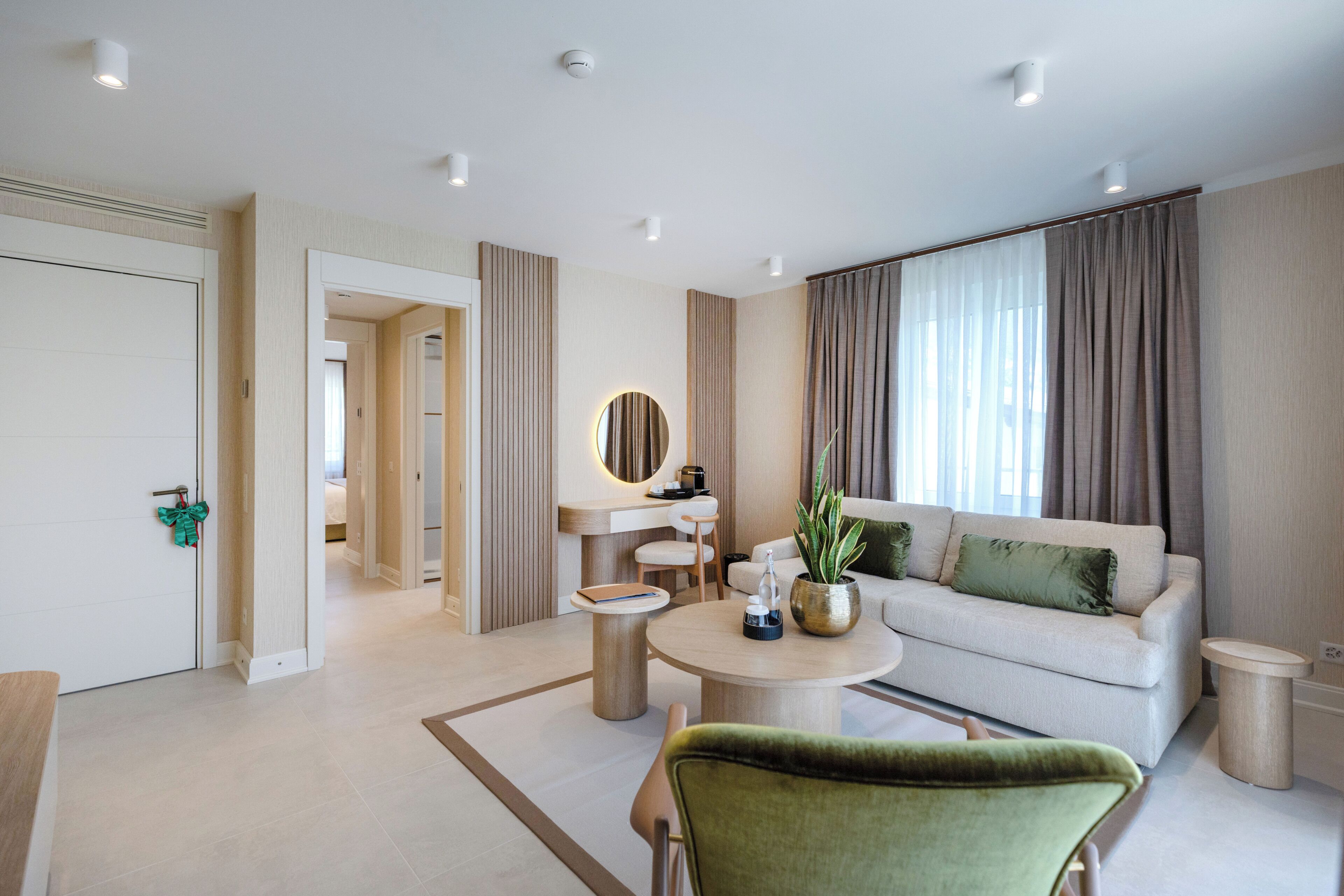 Ein modernes Wohnzimmer mit einer Couch, einem Couchtisch und einem grünen Sessel. Eine Tür führt in einen anderen Raum, und ein Fenster ist mit Vorhängen dekoriert.