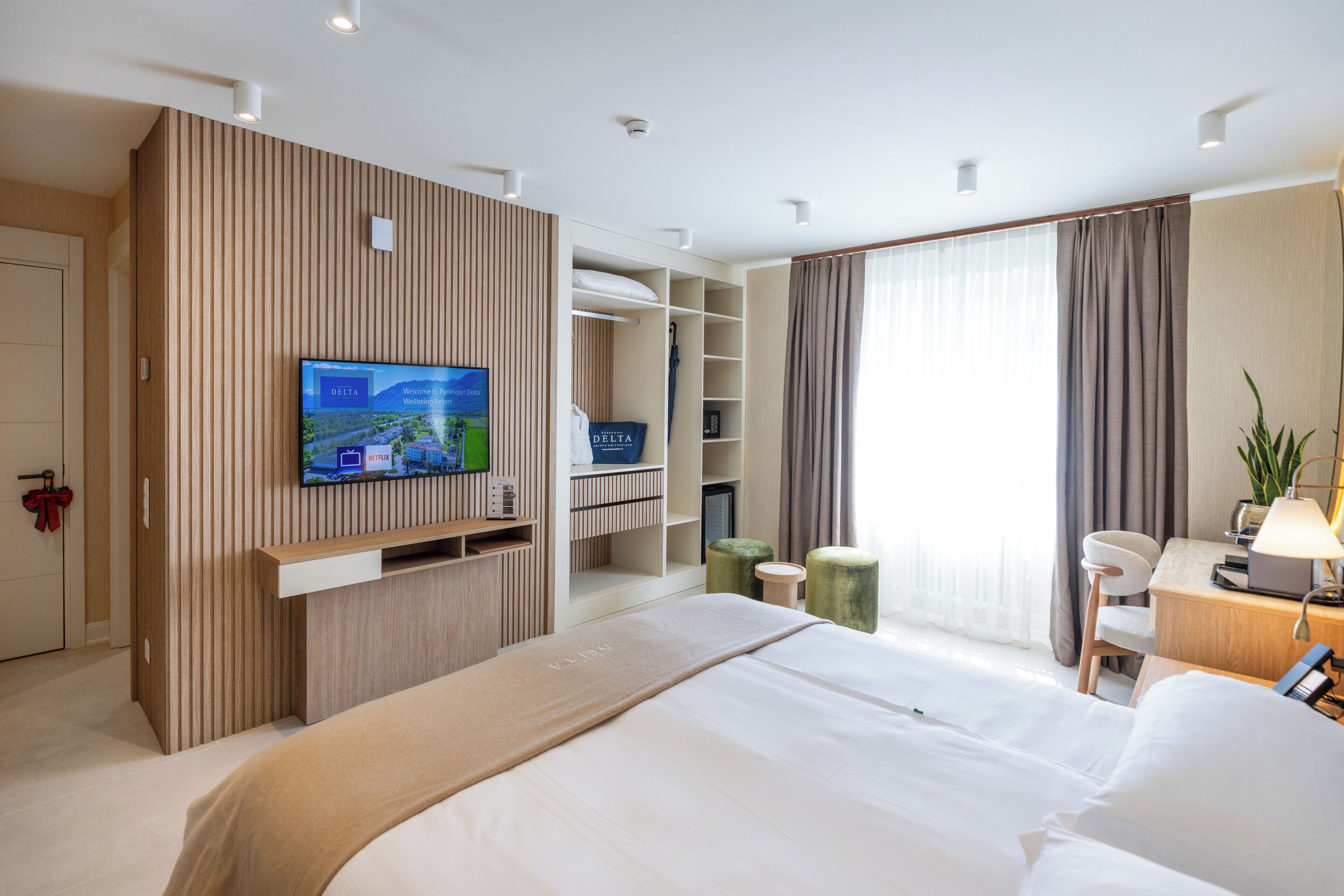 Habitación de hotel con cama, televisor montado en una pared de paneles de madera, escritorio con lámpara y ventana con cortinas.