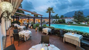 An outdoor dining area with tables set for two, overlooking a pool and a mountainous landscape. - Villa Delta – Parkhotel Delta (Ascona)