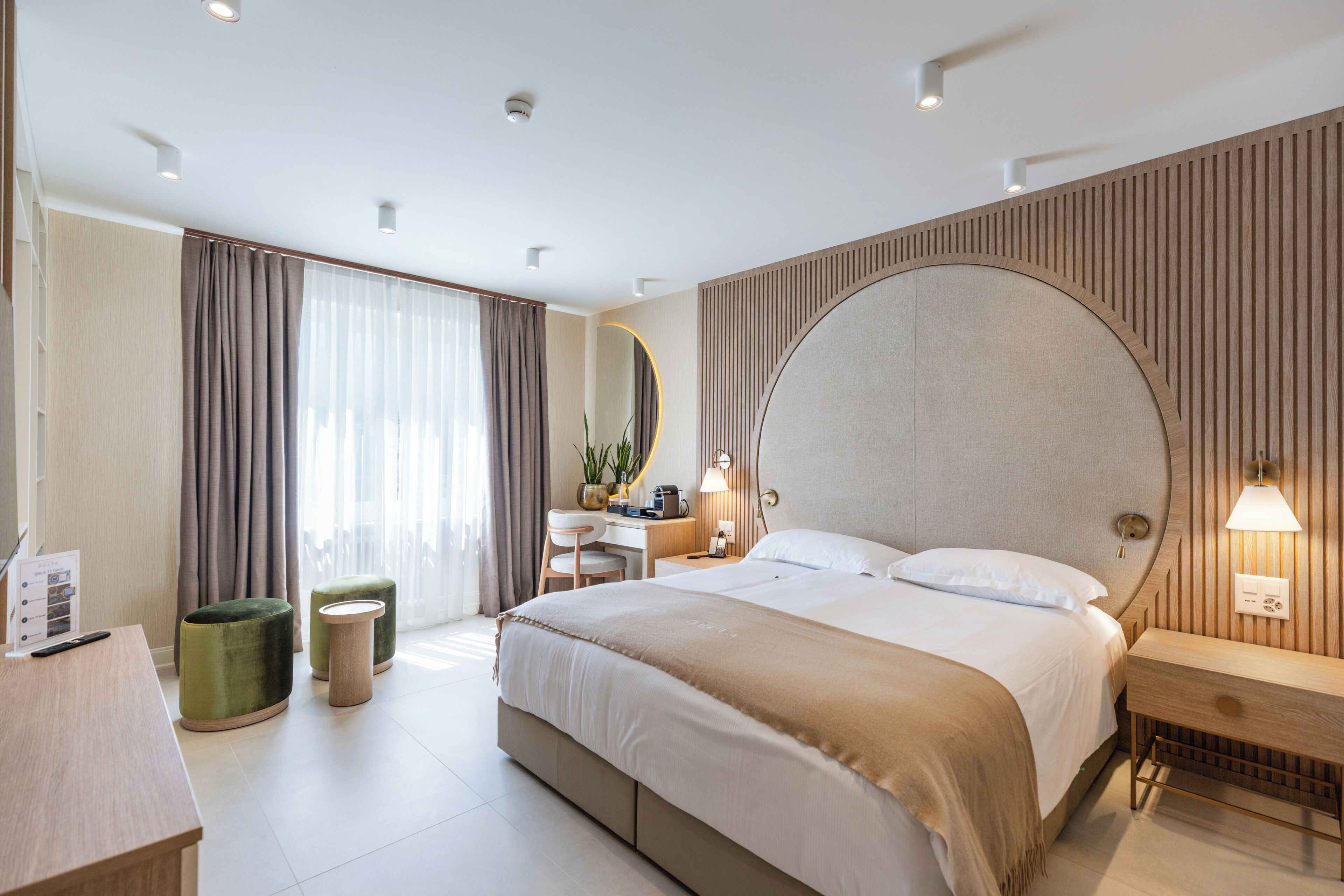 A modern hotel room with a large bed, a desk, a chair, a mirror, and a bedside table with a lamp.