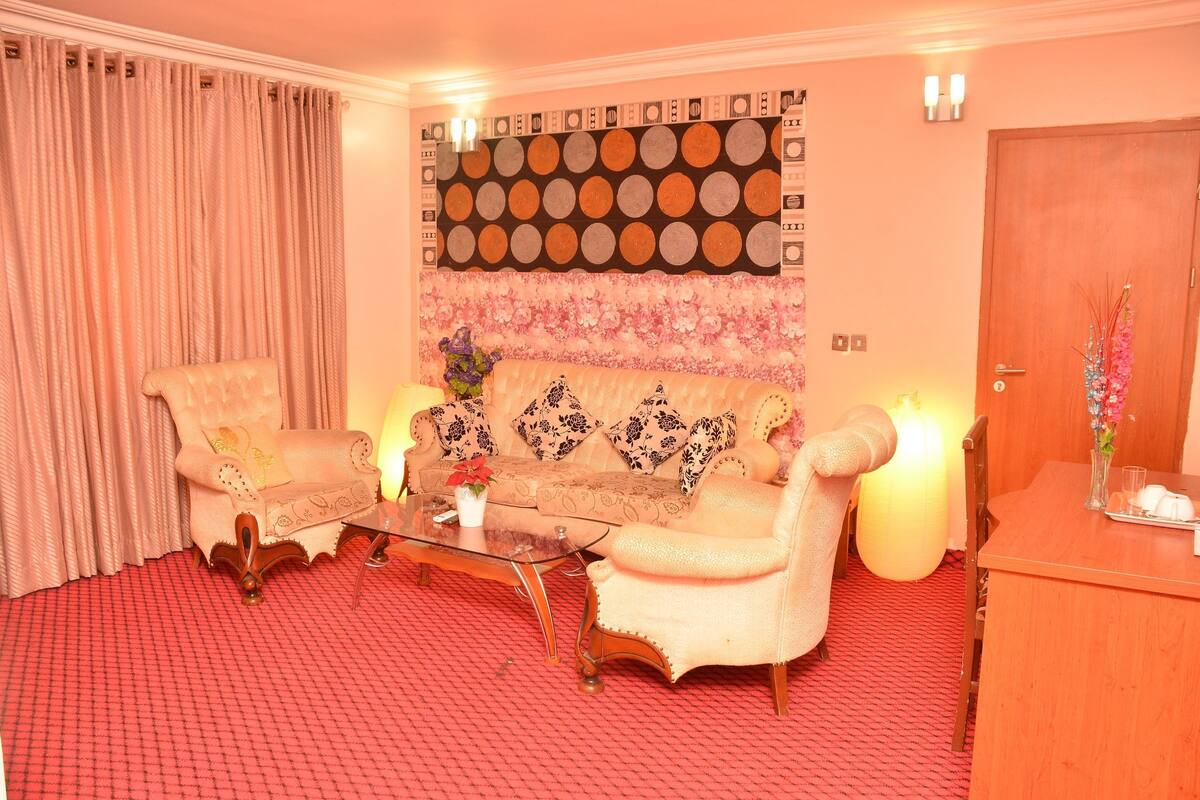 A room with a sofa, two armchairs, a glass coffee table, and a wooden side table with a vase of flowers.