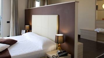 A modern hotel room with a large bed, bedside lamps, a small table, and a view of the city and sea.