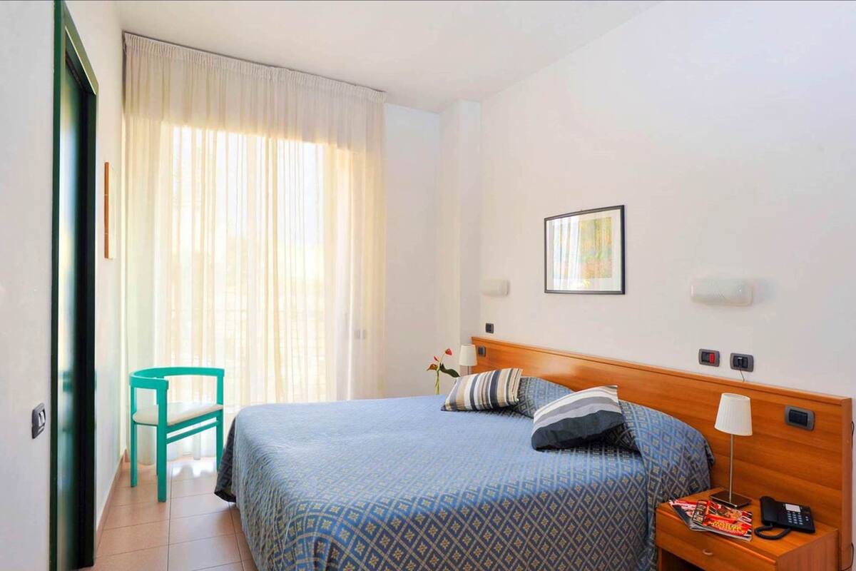 Habitación de hotel con una cama, una mesita de noche con lámpara, una silla y un ventanal con cortinas.